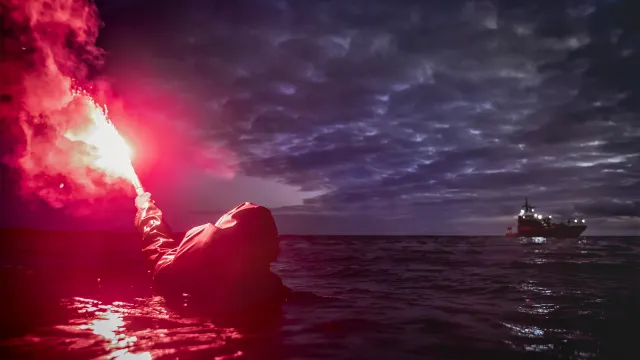 Person i vattnet avfyrar ett nödbloss. I bakgrunden syns en sjöräddningsbåt